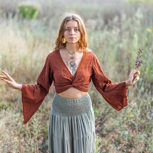 K&ouml;nnte beinhalten: Eine Frau tr&auml;gt ein rostfarbenes, lang&auml;rmeliges, bauchfreies Oberteil mit einem Twist-Detail vorne. Sie tr&auml;gt einen salbeigr&uuml;nen, mehrlagigen Rock mit einem Smok-Bund. Sie steht in einem Feld mit hohem Gras und h&auml;lt einen Zweig getrockneter Blumen.