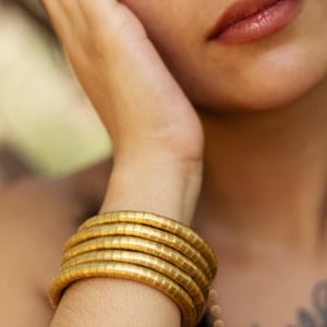 Könnte beinhalten: Ein Satz von fünf goldfarbenen, segmentierten Armbändern umschließt ein Handgelenk. Die Armbänder bestehen aus kleinen, verbundenen zylindrischen Teilen. Der Hautton ist hell, und der Hintergrund ist verschwommen, was auf eine Umgebung im Freien hindeutet. Die Armbänder sind der Mittelpunkt.