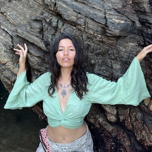 May include: A woman wearing a light green, long-sleeved, knotted crop top with bell sleeves. She is standing in front of a large, textured rock formation.