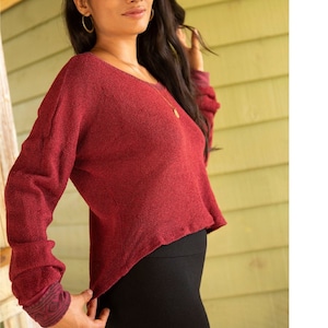 Puede incluir: Una mujer lleva un suéter color granate con un dobladillo alto-bajo y leggings negros. El suéter tiene una textura sutil y un pequeño detalle decorativo en el puño.