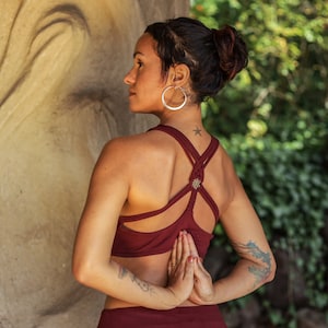 Puede incluir: Una mujer con un mo&ntilde;o en el pelo lleva un top de yoga rojo con un dise&ntilde;o de espalda cruzada. Est&aacute; de pie con las manos juntas frente a su pecho.