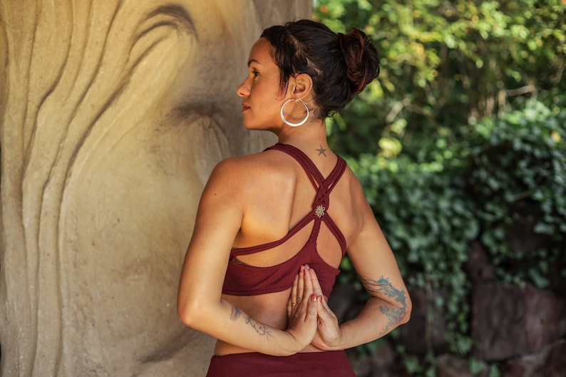 Puede incluir: Una mujer con un mo&ntilde;o en el pelo lleva un top de yoga rojo con un dise&ntilde;o de espalda cruzada. Est&aacute; de pie con las manos juntas frente a su pecho.