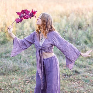 K&ouml;nnte beinhalten: Eine Frau tr&auml;gt ein locker sitzendes Oberteil und eine Hose in Lila mit einer Schleife in der Taille. Sie h&auml;lt einen Zweig mit roten Bl&auml;ttern und l&auml;chelt.