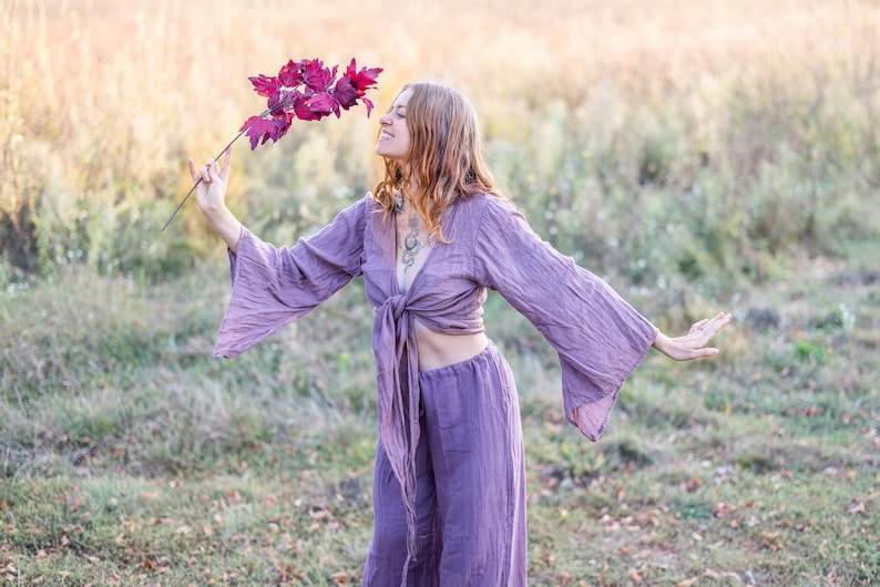 K&ouml;nnte beinhalten: Eine Frau tr&auml;gt ein locker sitzendes Oberteil und eine Hose in Lila mit einer Schleife in der Taille. Sie h&auml;lt einen Zweig mit roten Bl&auml;ttern und l&auml;chelt.