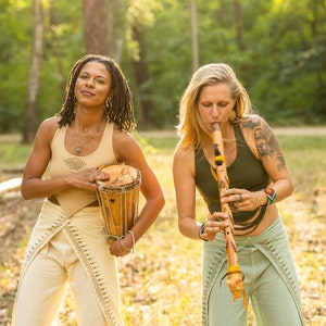 May include: Two people playing music outdoors. One person is playing a wooden flute, while the other is holding a small wooden drum. Both are wearing loose-fitting trousers and tank tops.