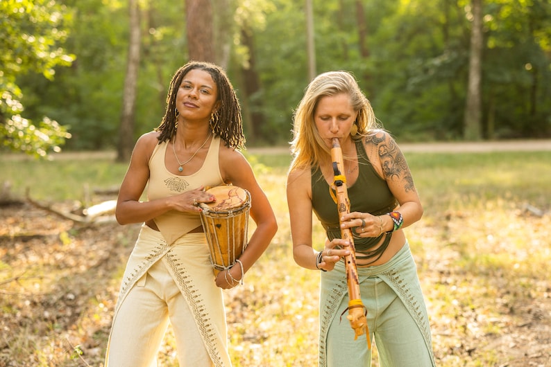 May include: Two people playing music outdoors. One person is playing a wooden flute, while the other is holding a small wooden drum. Both are wearing loose-fitting trousers and tank tops.