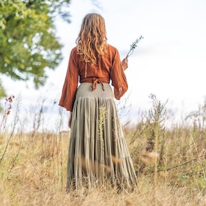 K&ouml;nnte beinhalten: Eine Frau tr&auml;gt einen langen, flie&szlig;enden Rock mit einem rostfarbenen Oberteil und einem braunen G&uuml;rtel. Sie steht in einem Feld mit hohem Gras und h&auml;lt einen kleinen Zweig mit Blumen.