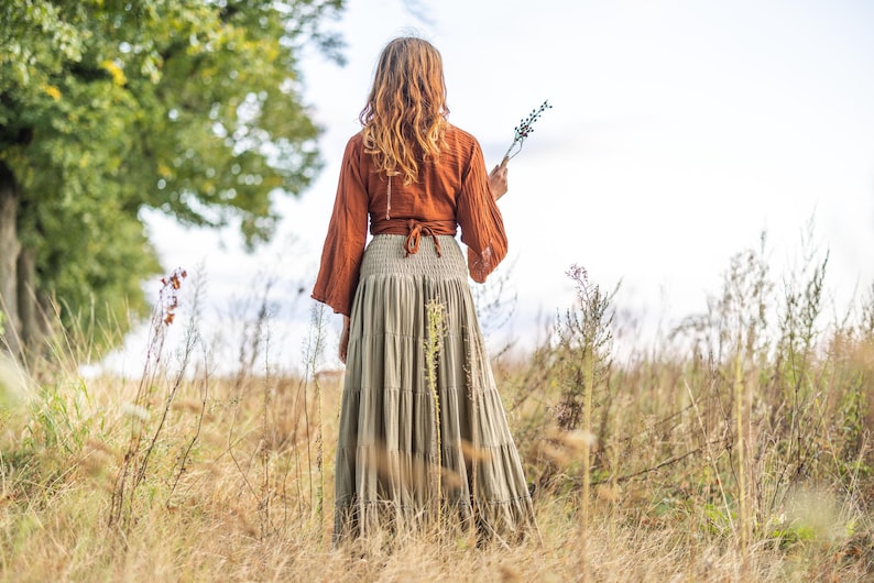 K&ouml;nnte beinhalten: Eine Frau tr&auml;gt einen langen, flie&szlig;enden Rock mit einem rostfarbenen Oberteil und einem braunen G&uuml;rtel. Sie steht in einem Feld mit hohem Gras und h&auml;lt einen kleinen Zweig mit Blumen.