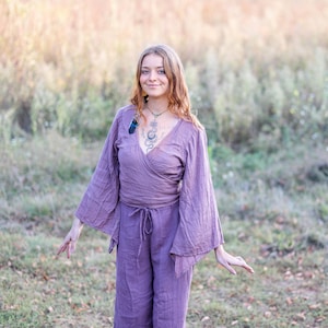 K&ouml;nnte beinhalten: Eine Frau tr&auml;gt ein lang&auml;rmliges, violettes Wickeltop mit einer Schleife in der Taille. Das Oberteil hat weite Glocken&auml;rmel und einen tiefen V-Ausschnitt. Sie steht auf einem Feld mit hohem Gras.