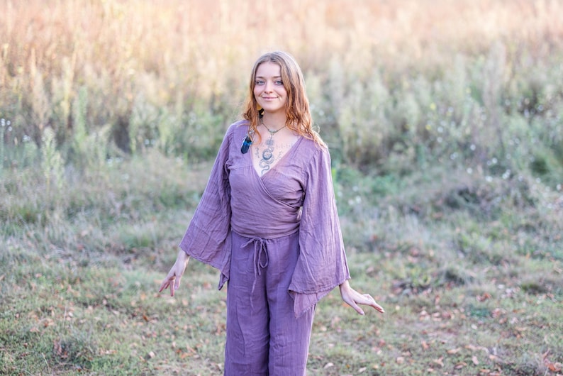 K&ouml;nnte beinhalten: Eine Frau tr&auml;gt ein lang&auml;rmliges, violettes Wickeltop mit einer Schleife in der Taille. Das Oberteil hat weite Glocken&auml;rmel und einen tiefen V-Ausschnitt. Sie steht auf einem Feld mit hohem Gras.