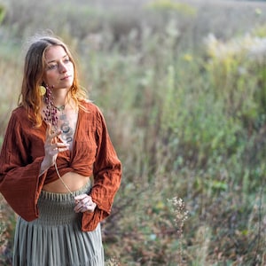 K&ouml;nnte beinhalten: Eine Frau tr&auml;gt ein locker sitzendes, lang&auml;rmliges Oberteil in Rostfarbe mit einem Knoten vorne und einen grauen Rock. Sie steht in einem Feld mit hohem Gras und h&auml;lt einen getrockneten Blumenst&auml;ngel.