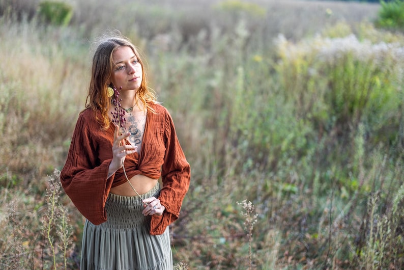 K&ouml;nnte beinhalten: Eine Frau tr&auml;gt ein locker sitzendes, lang&auml;rmliges Oberteil in Rostfarbe mit einem Knoten vorne und einen grauen Rock. Sie steht in einem Feld mit hohem Gras und h&auml;lt einen getrockneten Blumenst&auml;ngel.