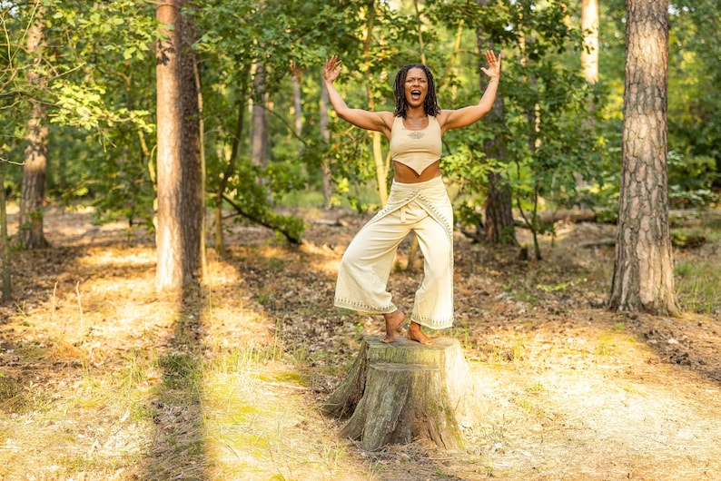 May include: A woman wearing a beige top and trousers stands on a tree stump in a forest. She is raising her arms and shouting.