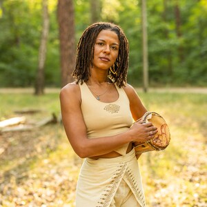 May include: A woman with dreadlocks is holding a brown wooden drum in a woodland setting. She is wearing a beige tank top with a design on the front and a beige skirt with a pattern.