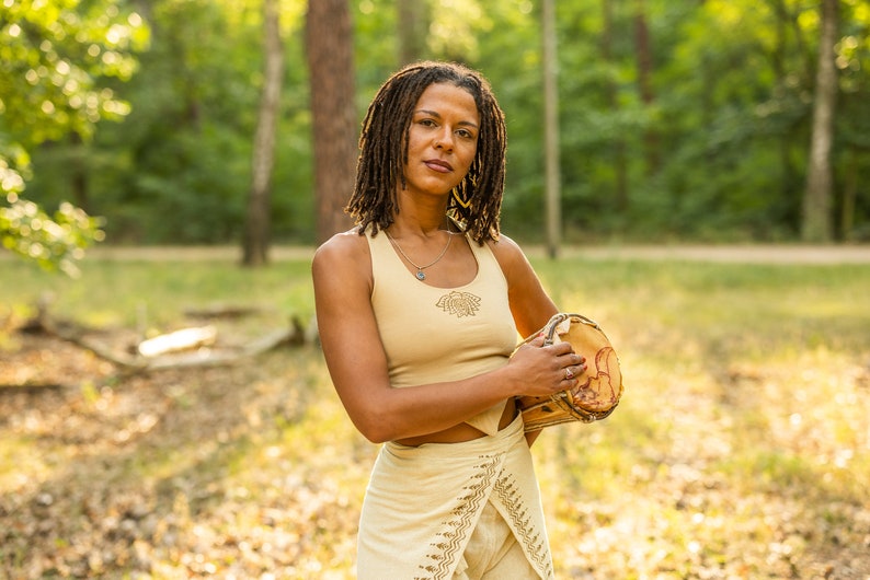 May include: A woman with dreadlocks is holding a brown wooden drum in a woodland setting. She is wearing a beige tank top with a design on the front and a beige skirt with a pattern.