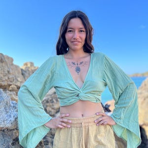 K&ouml;nnte beinhalten: Eine Frau tr&auml;gt ein hellgr&uuml;nes, lang&auml;rmliges, geknotetes Crop-Top mit Glocken&auml;rmeln und eine beige, hochgeschlossene Shorts mit Fransen am Saum.