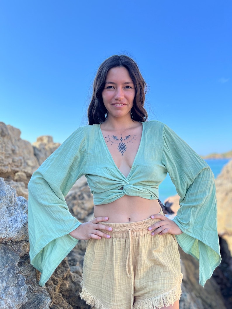 K&ouml;nnte beinhalten: Eine Frau tr&auml;gt ein hellgr&uuml;nes, lang&auml;rmliges, geknotetes Crop-Top mit Glocken&auml;rmeln und eine beige, hochgeschlossene Shorts mit Fransen am Saum.