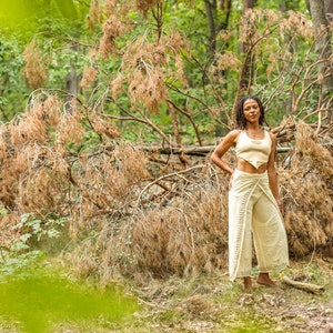 May include: A woman wearing a white crop top and white trousers stands in a woodland setting. The trousers have a wrap design and are tied at the waist. The woman is standing in front of a fallen tree with brown branches.