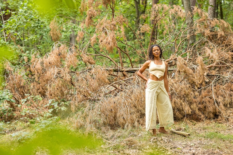 May include: A woman wearing a white crop top and white trousers stands in a woodland setting. The trousers have a wrap design and are tied at the waist. The woman is standing in front of a fallen tree with brown branches.