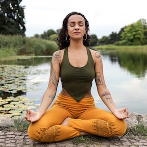 Puede incluir: Una persona sentada en posici&oacute;n de loto en un camino de piedra junto a un estanque, con un top verde y pantalones de yoga amarillo mostaza con un patr&oacute;n geom&eacute;trico. La persona tiene tatuajes en los brazos y est&aacute; meditando con los ojos cerrados.
