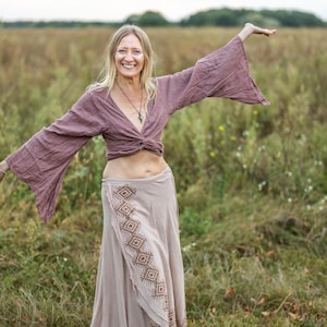 K&ouml;nnte beinhalten: Eine Frau tr&auml;gt ein langes, flie&szlig;endes, lila Oberteil mit einem braunen und wei&szlig;en gemusterten Rock. Sie steht auf einem Feld mit hohem Gras und l&auml;chelt.