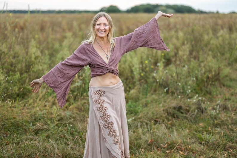 K&ouml;nnte beinhalten: Eine Frau tr&auml;gt ein langes, flie&szlig;endes, lila Oberteil mit einem braunen und wei&szlig;en gemusterten Rock. Sie steht auf einem Feld mit hohem Gras und l&auml;chelt.