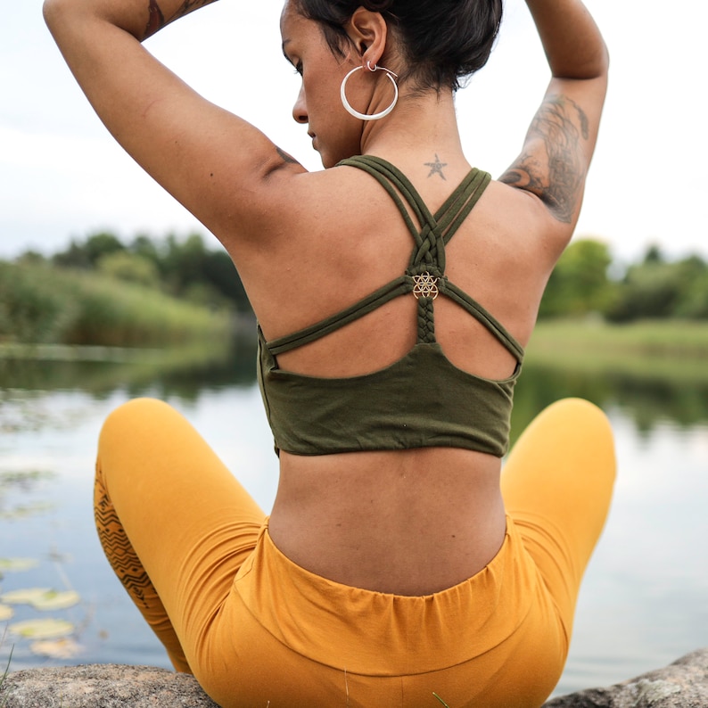 Puede incluir: Una mujer con un top de yoga verde con un detalle de espalda trenzada y un dije dorado. Tambi&eacute;n lleva leggings de color mostaza con un patr&oacute;n geom&eacute;trico.
