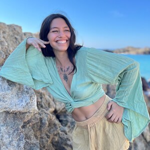 K&ouml;nnte beinhalten: Eine Frau tr&auml;gt eine lockere, lang&auml;rmelige Bluse in Hellgr&uuml;n mit einem Knoten vorne und eine hellbeige, kurze Hose aus Leinen. Sie l&auml;chelt und posiert mit ihrer Hand in der N&auml;he ihres Gesichts.