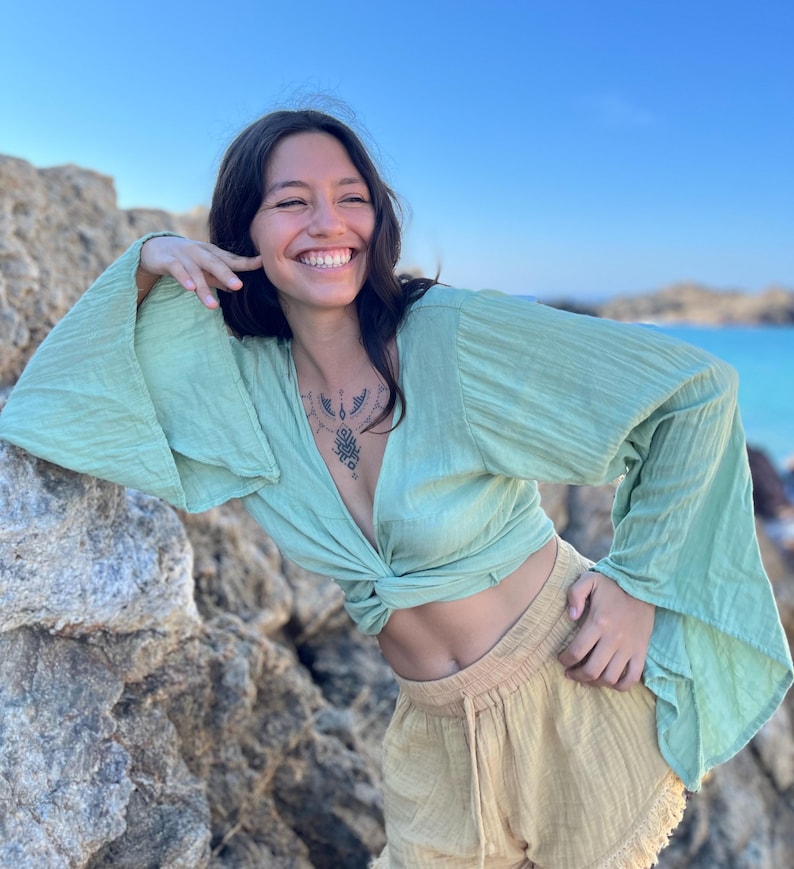 K&ouml;nnte beinhalten: Eine Frau tr&auml;gt eine lockere, lang&auml;rmelige Bluse in Hellgr&uuml;n mit einem Knoten vorne und eine hellbeige, kurze Hose aus Leinen. Sie l&auml;chelt und posiert mit ihrer Hand in der N&auml;he ihres Gesichts.