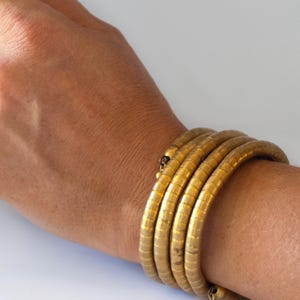 May include: A gold-colored, coiled bracelet wraps around a wrist. The bracelet is made of multiple, connected segments, creating a flexible, textured appearance. A silver ring is visible on a finger.