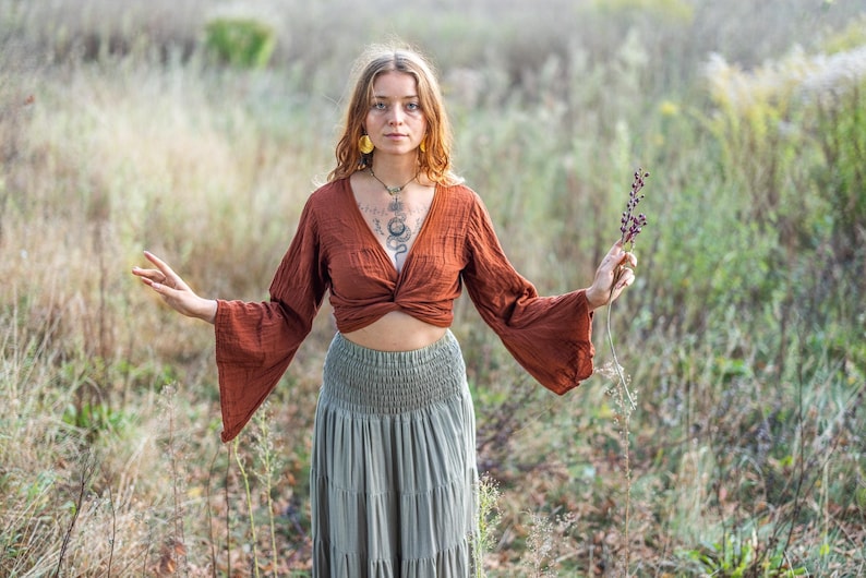 K&ouml;nnte beinhalten: Eine Frau tr&auml;gt ein rostfarbenes, lang&auml;rmeliges, bauchfreies Oberteil mit einem Twist-Detail vorne. Sie tr&auml;gt einen salbeigr&uuml;nen, mehrlagigen Rock mit einem Smok-Bund. Sie steht in einem Feld mit hohem Gras und h&auml;lt einen Zweig getrockneter Blumen.