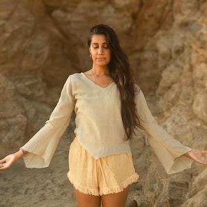 Op de afbeelding: Een persoon draagt een beige top met lange mouwen en uitlopende mouwen en een bijpassende short met een gefranjerde zoom. De outfit is licht van kleur en lijkt gemaakt van een zachte stof. De achtergrond is een natuurlijke rotsformatie.
