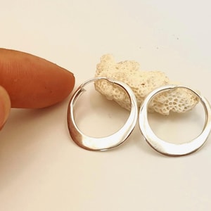 May include: A pair of silver-coloured, open-circle earrings. The earrings have a smooth, polished finish and a slightly irregular shape. They are displayed on a white surface next to a piece of coral.
