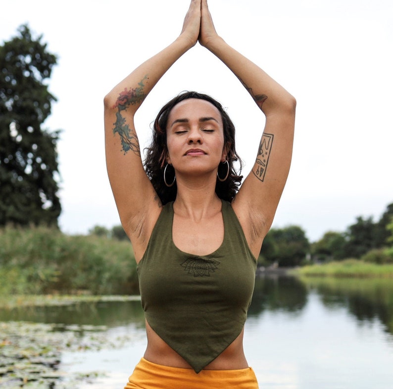 Puede incluir: Una mujer con cabello oscuro y un tatuaje en el brazo lleva un top verde sin mangas con un dobladillo triangular y una falda amarilla. Est&aacute; de pie con los brazos levantados en una postura de yoga, con los ojos cerrados, frente a un cuerpo de agua.