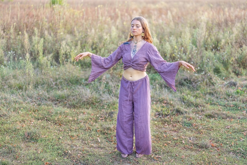 K&ouml;nnte beinhalten: Eine Frau tr&auml;gt ein lilafarbenes, lang&auml;rmeliges, locker sitzendes Oberteil mit einem geknoteten Vorderteil und eine passende weite Hose. Sie steht in einem Feld mit hohem Gras und hat die Arme ausgestreckt.