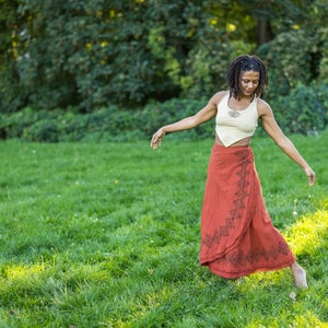 May include: A woman with long dreadlocks is wearing a white crop top and a long red wrap skirt with black geometric patterns. She is standing in a grassy field with trees in the background.