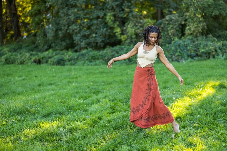May include: A woman with long dreadlocks is wearing a white crop top and a long red wrap skirt with black geometric patterns. She is standing in a grassy field with trees in the background.