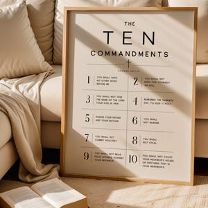 May include: A framed print featuring "THE TEN COMMANDMENTS" in a minimalist design. The print has a light beige background with black text, set within a wooden frame. The image is displayed against a neutral backdrop.