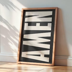 Könnte beinhalten: Ein gerahmter Kunstdruck mit dem Wort "AMEN" in großen, weißen, fetten Buchstaben auf schwarzem Hintergrund. Der Rahmen ist aus hellem Holz. Das Kunstwerk lehnt an einer weißen Wand.