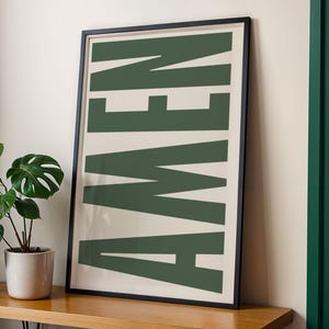 Könnte beinhalten: Ein gerahmter Kunstdruck mit dem Wort "AMEN" in großen, dunkelgrünen Blockbuchstaben auf cremefarbenem Hintergrund. Der Druck befindet sich in einem schwarzen Rahmen und lehnt an einer Wand. Eine Topfpflanze steht in der Nähe auf einer Holzoberfläche.