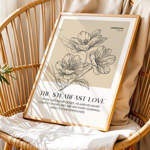 May include: A framed art print featuring a floral illustration in black and white. The print includes the text "THE STEADFAST LOVE" and a quote from Lamentations. The artwork is displayed on a wicker chair.