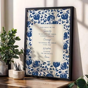 May include: Framed wall art featuring a floral pattern in blue and white. The artwork includes the text "God grant me the Serenity..." in a central cream-colored rectangle. The frame is black, and the art is displayed on a wooden surface.