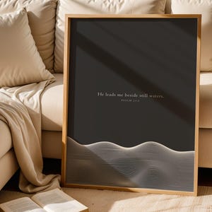May include: A framed art print with a dark gray background and a minimalist white wave design at the bottom. The text "He leads me beside still waters. Psalm 23:2" is printed in a light color at the top. The frame is made of light-colored wood.