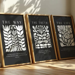 Puede incluir: Tres impresiones artísticas enmarcadas con marcos dorados. Cada impresión tiene un fondo negro con diseños florales blancos y texto. El texto de las impresiones dice "THE WAY", "THE TRUTH" y "THE LIFE". Las impresiones están apoyadas contra una pared.