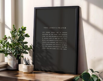May include: Black framed print featuring "The Lord's Prayer" in white text. The print is displayed on a wooden surface, next to a potted plant and a pencil holder. The overall aesthetic is minimalist and modern.