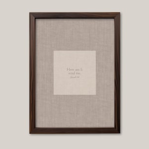 May include: Framed artwork featuring a linen-textured background with a central square containing the text "Here am I; send me." The frame is a dark brown wood. The artwork is displayed against a neutral-colored wall.