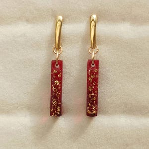 Peut inclure: Une paire de boucles d'oreilles dorées avec un pendentif rectangulaire en résine rouge. Le pendentif est incrusté de paillettes dorées. Les boucles d'oreilles ont un design simple et élégant.