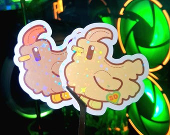 May include: Two holographic stickers featuring a cartoon chicken design. The stickers have a light brown and yellow color scheme with a glittery, star-like effect. Each sticker has a small heart with the letters "SD".