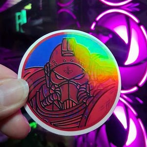 May include: A round sticker featuring a red and black illustration of a helmeted figure. The sticker has a blue background and a rainbow-colored iridescent effect on the right side. The sticker is held by a person's hand.