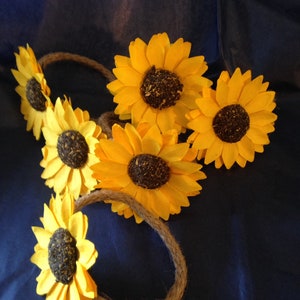 May include: A collection of sunflower headbands with bright yellow petals and dark brown centers. The sunflowers are attached to a natural-colored rope band, creating a rustic and summery aesthetic. The headbands are arranged on a dark blue background.